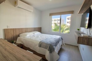 A bedroom with a bed, a desk, a TV, and a window with a view of buildings and palm trees.