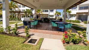 Un patio exterior con mesas y sillas, palmeras y una zona cubierta con ventiladores de techo.