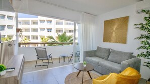 A modern living room with a grey sofa, a yellow armchair, a small round table, and a balcony with a view of a building and greenery.