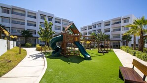A playground with a slide and swings, surrounded by a well-maintained lawn and a building in the background.