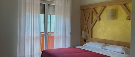 Un dormitorio con estructura de cama de madera, cubrecama rojo, una mesa de madera y un balcón con cortinas blancas.