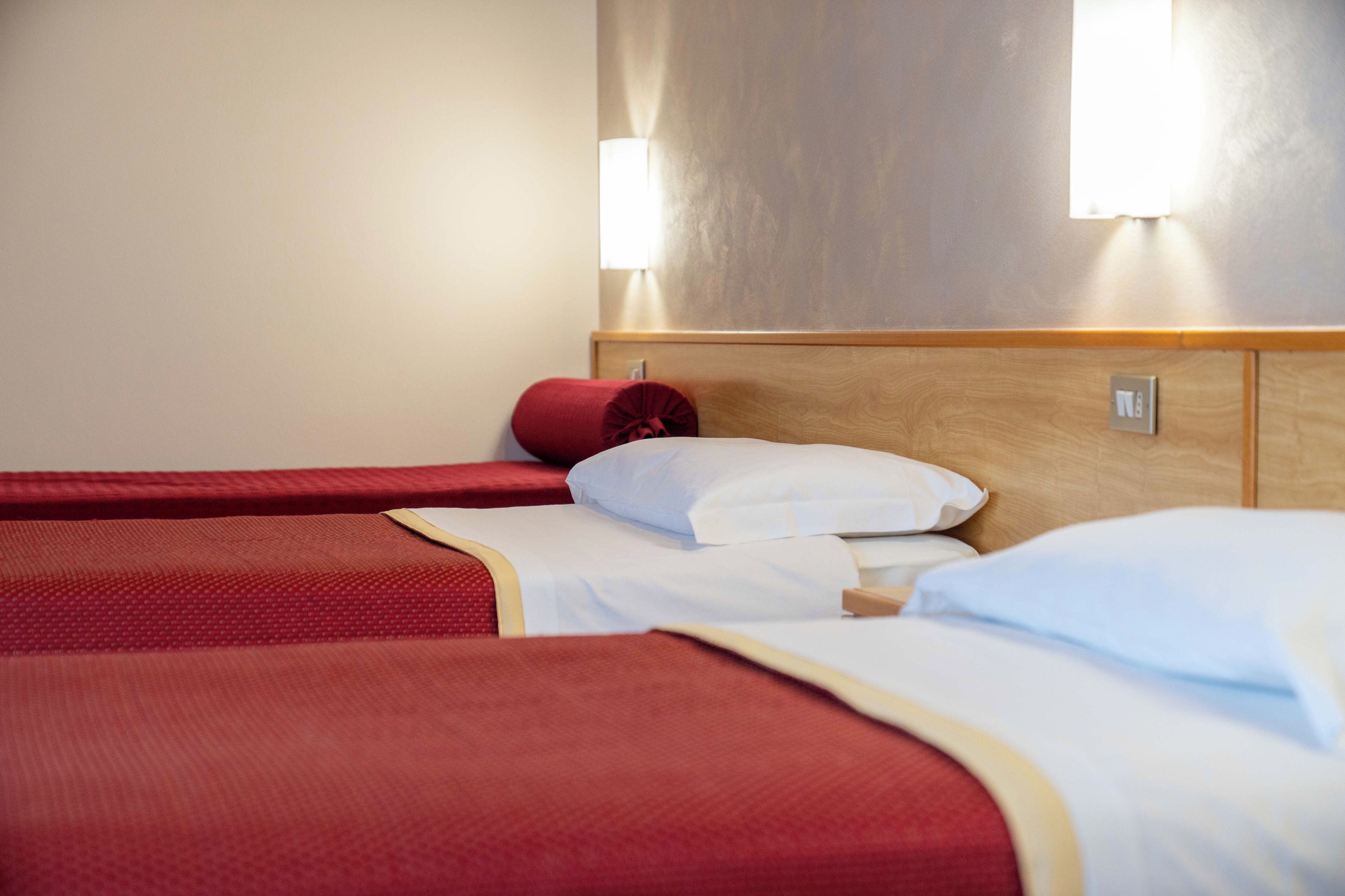 Habitación de hotel con dos camas, cada una con ropa de cama roja y almohadas blancas. Hay dos luces fijadas a la pared sobre las camas.