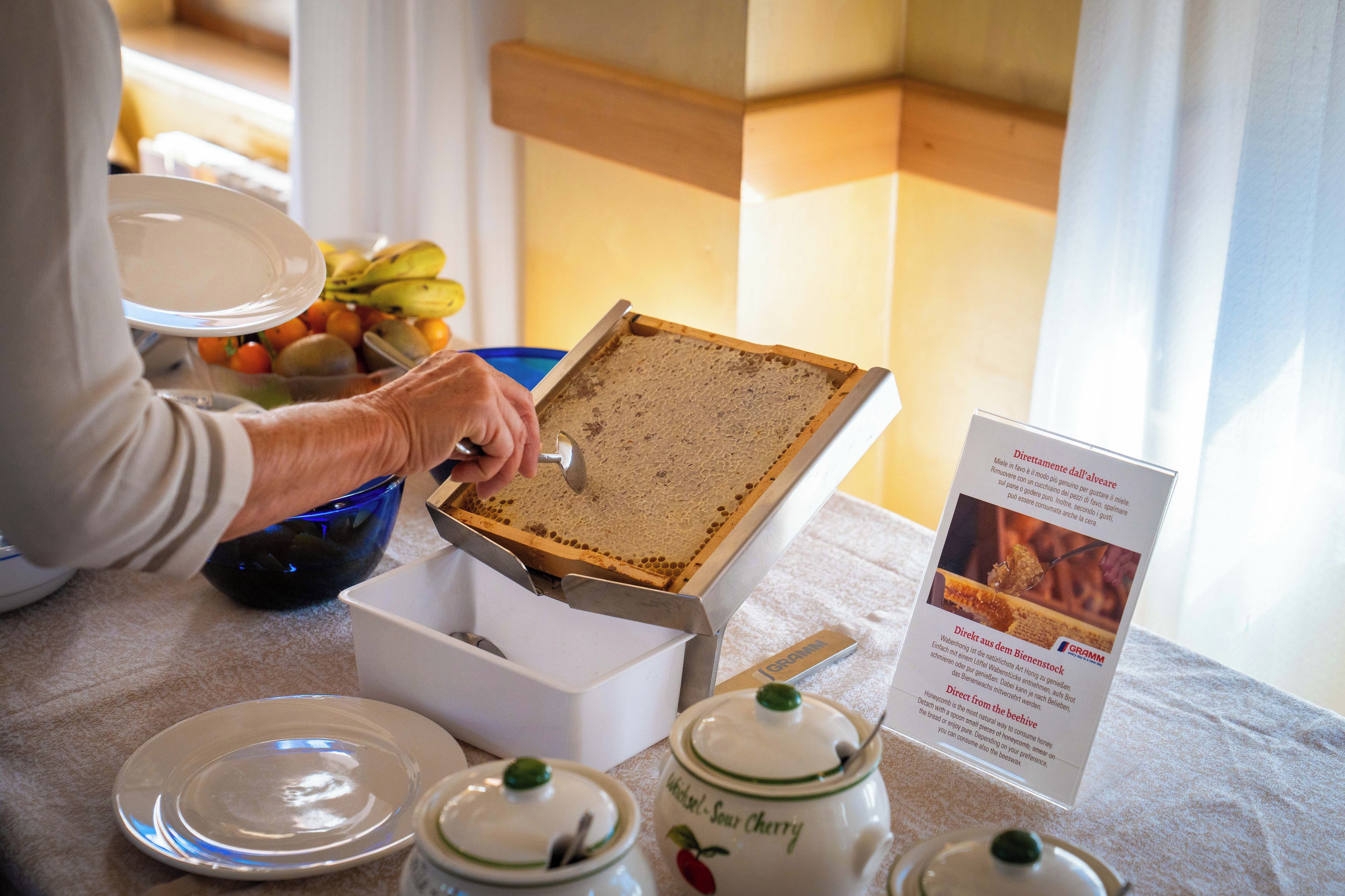 A person opening a honeycomb box, with a bowl of fruit and honey jars on the table.