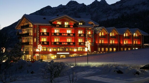 Um hotel grande, com vários andares, fachada vermelha e varandas, cercado por montanhas cobertas de neve.