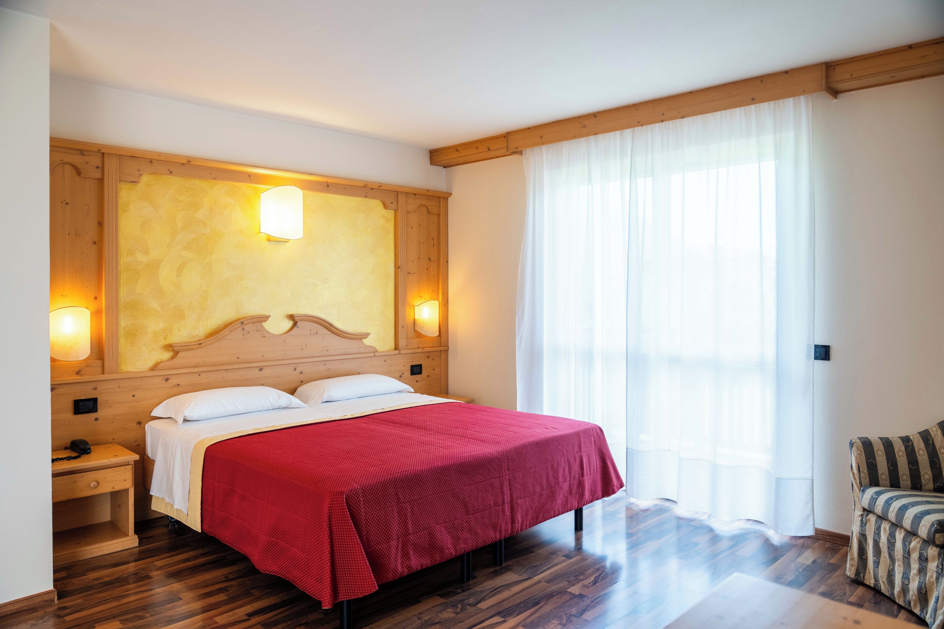 A hotel room with a wooden headboard, a red bedspread, a nightstand with a phone, and a window with sheer curtains.