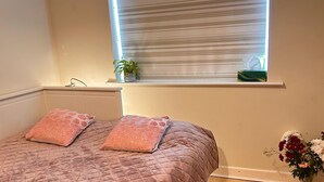 Une chambre à coucher avec un lit, une fenêtre avec des stores, une table de chevet avec une plante et une boîte de mouchoirs, et un vase avec des fleurs.