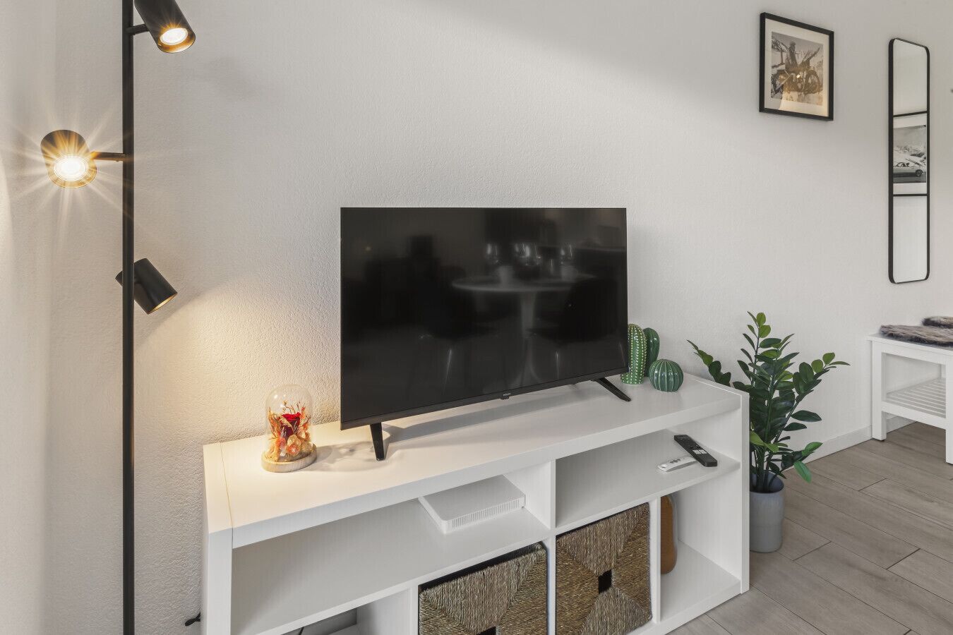 Una sala moderna con un televisor de pantalla plana sobre un mueble blanco, una lámpara de pie, un cactus y un cuadro en la pared.