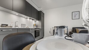 Une cuisine moderne avec des meubles de couleur sombre, un plan de travail blanc et un coin repas agrémenté d’une carafe en verre et d’assiettes.