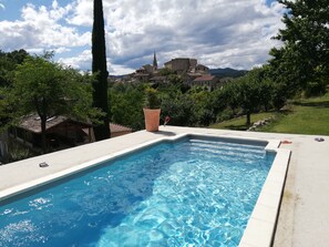 Une piscine à l’eau d’un bleu cristallin, entourée d’une terrasse en béton. Au fond, on aperçoit une ville avec ses bâtiments et le clocher d’une église.