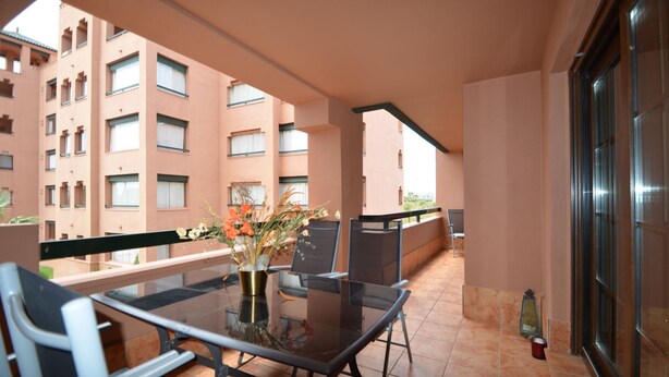 A balcony with a glass table and chairs, a vase with flowers, and a view of a building with multiple windows. - PLAYA VERDE 37 - Two Bedroom Apartment, Sleeps 5 (Punta del moral)