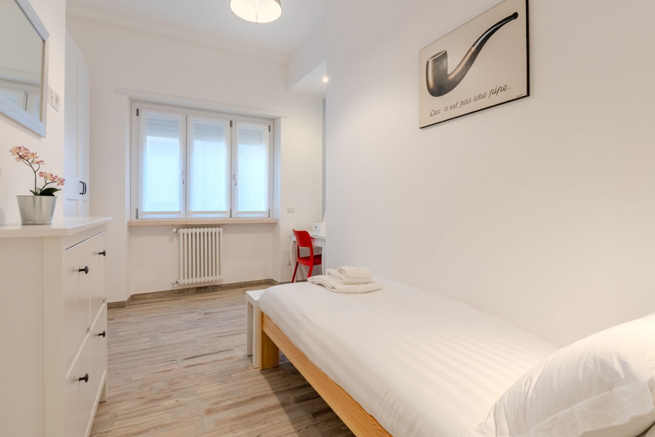 A modern bedroom with a wooden bed, a white dresser, a red chair, a window with blinds, and a radiator.