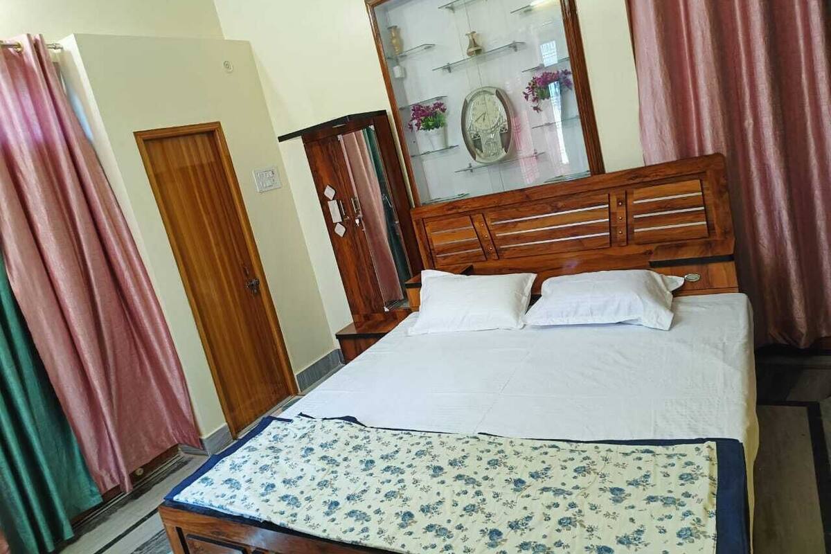 A bedroom with a wooden bed, white bedding, a floral-patterned throw, a wooden wardrobe, and a glass-fronted cabinet with decorative items.