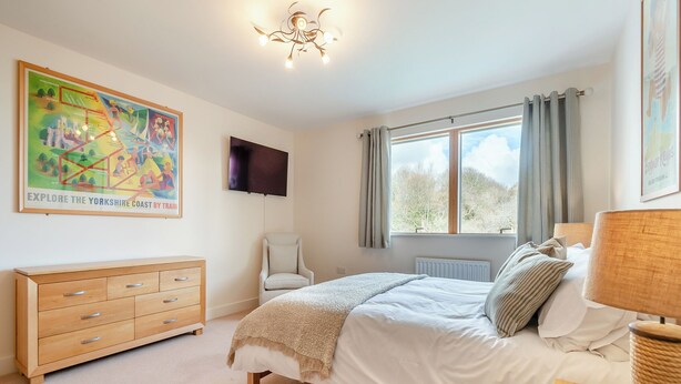 Un dormitorio con cama, una silla, tocador, televisor y ventana con cortinas.