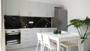 A modern kitchen with white cabinets, a black marble backsplash, and a stainless steel refrigerator. There is a dining table with white chairs and a potted plant.