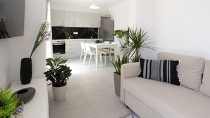 A modern living room with a beige sofa, a dining area with white chairs, and a kitchen with white cabinets and a black countertop.