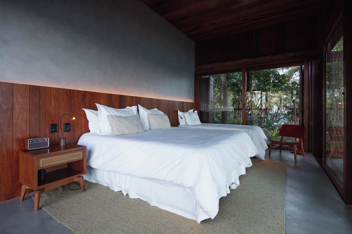 A bedroom with a large bed, wooden headboard, bedside table, and a view of the outdoors through large windows.