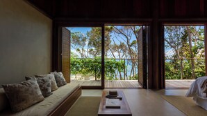 A room with a sofa, a coffee table, and a view of a wooden deck and trees. - Villa Toucan (Itacaré)