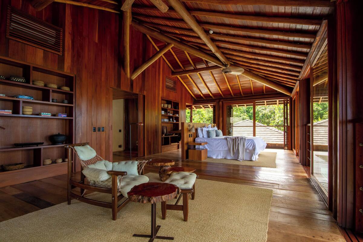 A spacious room with wooden walls and ceiling, a large bed, a seating area with a chair and ottoman, and a view of greenery outside.