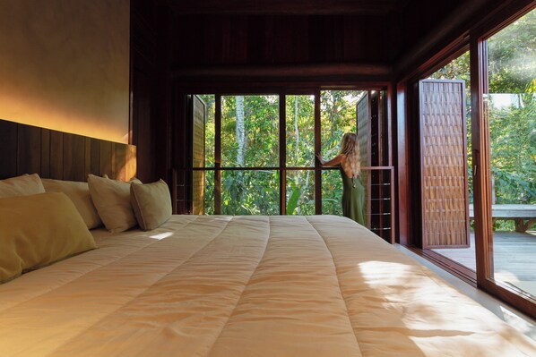 A large bed with a quilted cover, a wooden headboard, and a view of greenery through large windows. - Villa Toucan (Itacaré)