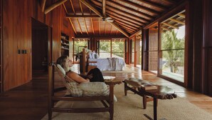 A spacious room with a high ceiling, wooden beams, a large bed, a sitting area with a chair and table, and large windows offering a view of palm trees. - Villa Toucan (Itacaré)