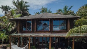 A wooden two-story building with large windows, a covered deck with lounge chairs, and a hammock. - Villa Toucan (Itacaré)