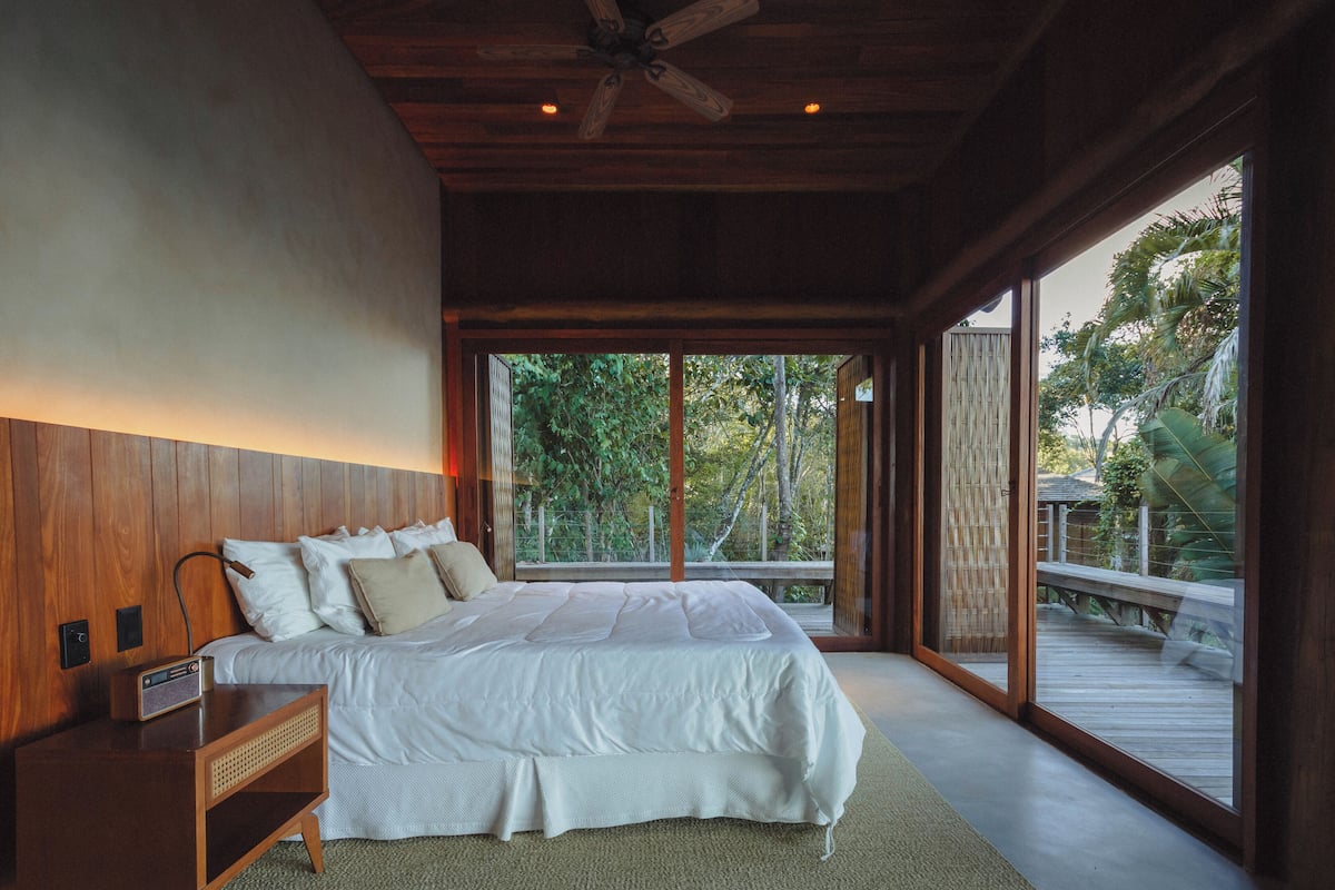 A bedroom with a large bed, wooden headboard, and a view of a wooden deck and greenery outside.