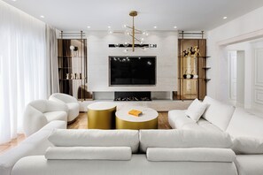 Ein modernes Wohnzimmer mit einem großen Flachbildfernseher, einer goldfarbenen Chandelüster und einer weißen Sofagarnitur.