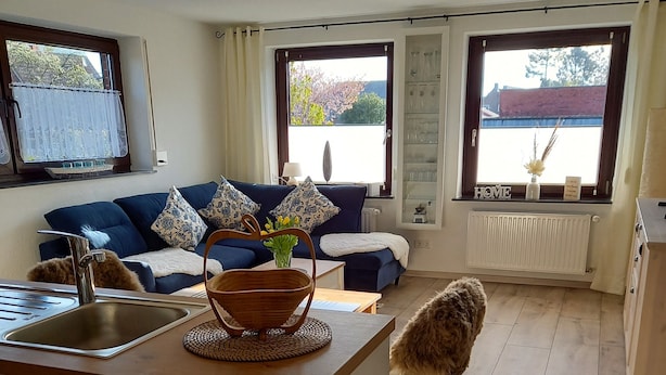 Una cocina moderna con encimera de madera, un fregadero y una zona de comedor con mesa y sillas. Se aprecia un sofá azul con cojines estampados al fondo.