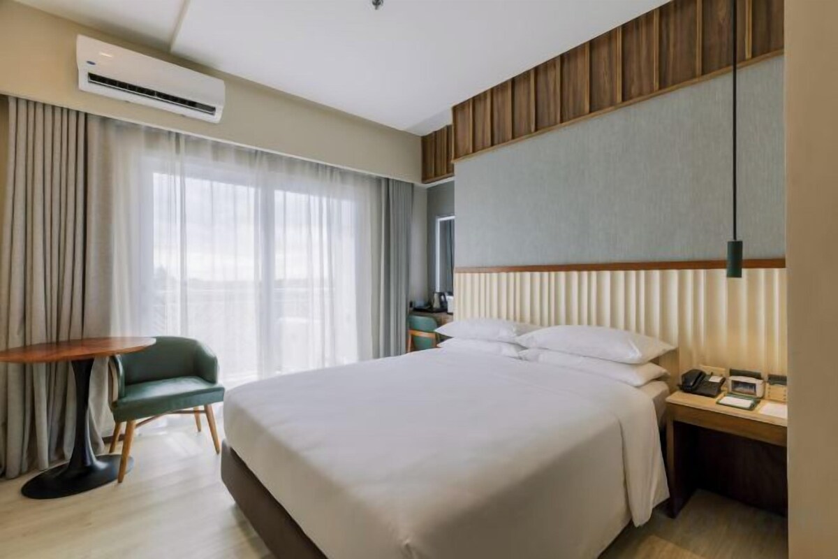 Habitación de hotel con una cama grande, un escritorio, una silla, una mesita de noche con lámpara y una ventana con cortinas.