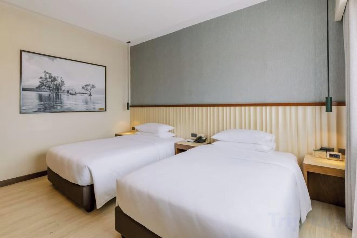 Habitación de hotel con dos camas individuales, una mesita de noche con lámpara, una mesita auxiliar y un cuadro en la pared.