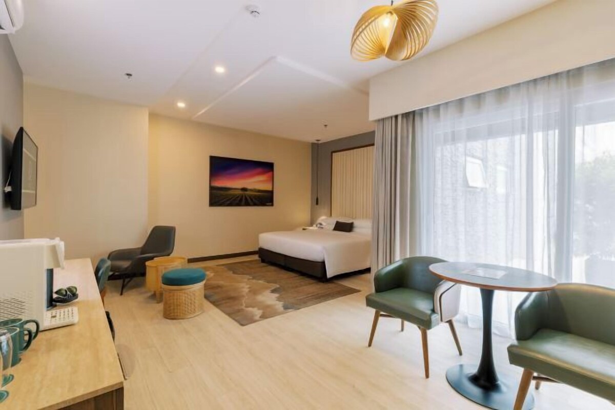 A modern hotel room with a bed, a desk with a computer, a chair, a round table, and a large window with curtains.