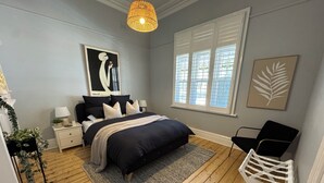 Une chambre à coucher comprenant un lit, des tables de chevet, une lampe, une fenêtre avec des stores, un luminaire suspendu et une œuvre d’art accrochée au mur.