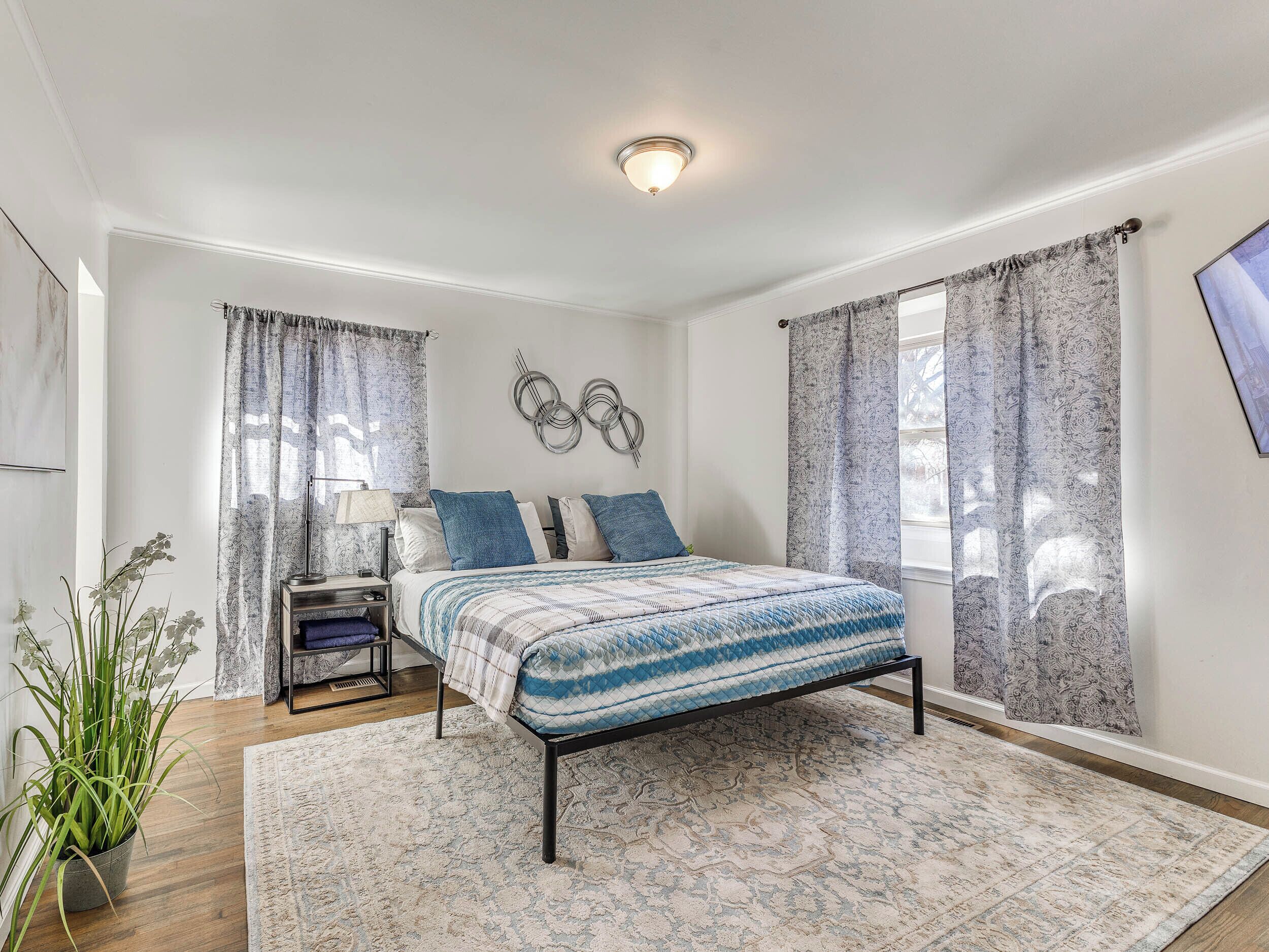 A bedroom with a bed, curtains, a wall art piece, a nightstand, a lamp, and a potted plant.