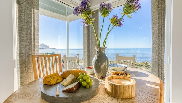 A table with cheese, grapes, and a candle, overlooking a beach with a clear view of the ocean. - Dihangfa - sleeps 6 guests  in 3 bedrooms (Nefyn)