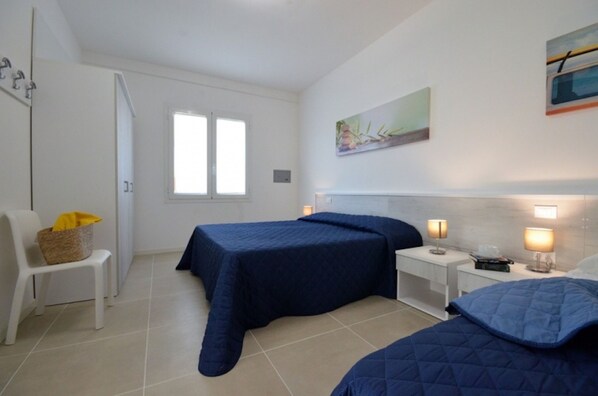 A bedroom with two beds, a white chair, a bedside table with lamps, and a window.
