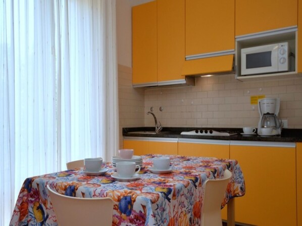 Eine kleine Küche mit gelben Schränken, einer Mikrowelle und einer Kaffeemaschine. Es gibt einen Tisch mit einer blumigen Tischdecke sowie weiße Tassen und Untertassen.