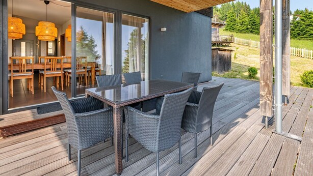 A covered outdoor dining area with a glass table and wicker chairs, overlooking a wooden deck and a forested area. - Beautiful apartment for 6 people with WIFI, TV, terrace and pets allowed (Frauenstein)