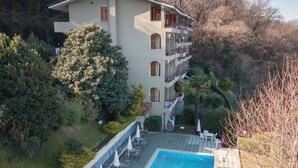Un hotel de varios pisos con piscina, mobiliario exterior y abundante vegetación.