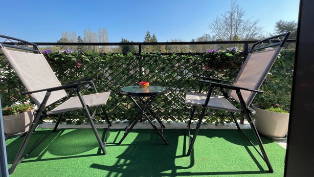 Ein Balkon mit zwei Stühlen und einem kleinen Tisch, umgeben von einem Gitterzaun und Grünpflanzen.