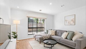 A living room with a grey sofa, a round coffee table, a TV, and a large window with a view of greenery.