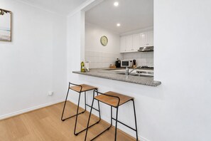 A modern kitchen with white cabinets, a granite countertop, a stainless steel sink, and a microwave. There are two bar stools with wooden seats and black metal frames. A clock is visible on the wall above the countertop. - Tullamarine Landing by Ready Set Stay (Tullamarine)