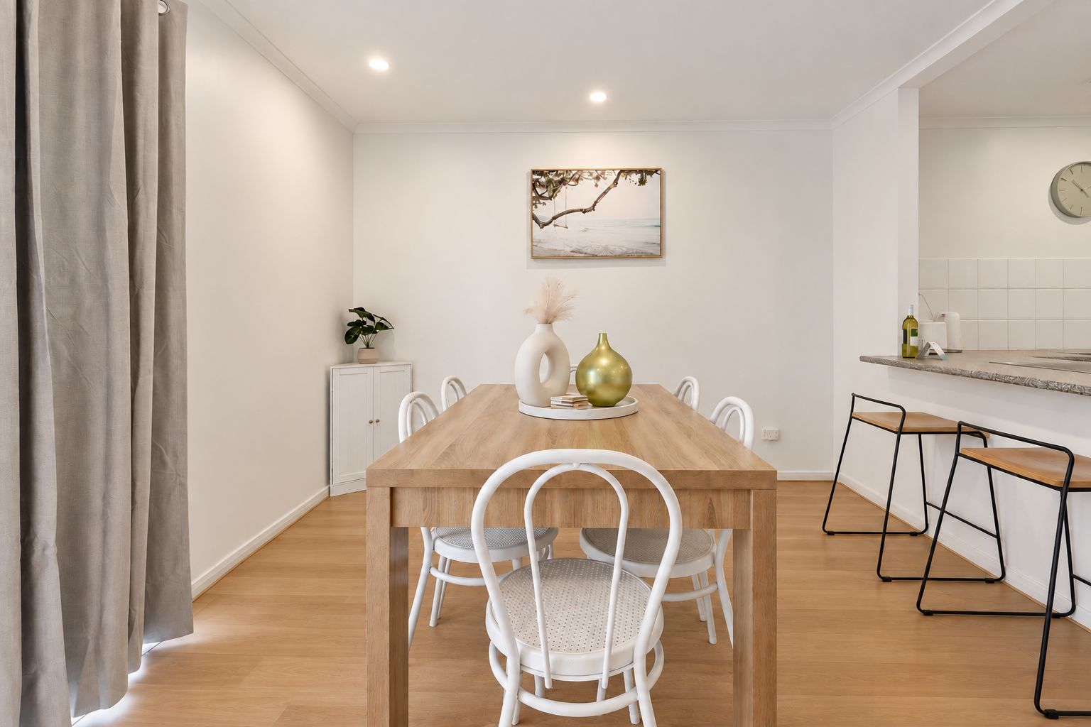 A dining area with a wooden table, white chairs, a small cabinet, a plant, and a painting on the wall.