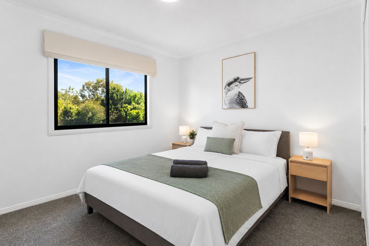 A neatly arranged bedroom with a bed, bedside tables, a window with a view, and a framed picture on the wall.