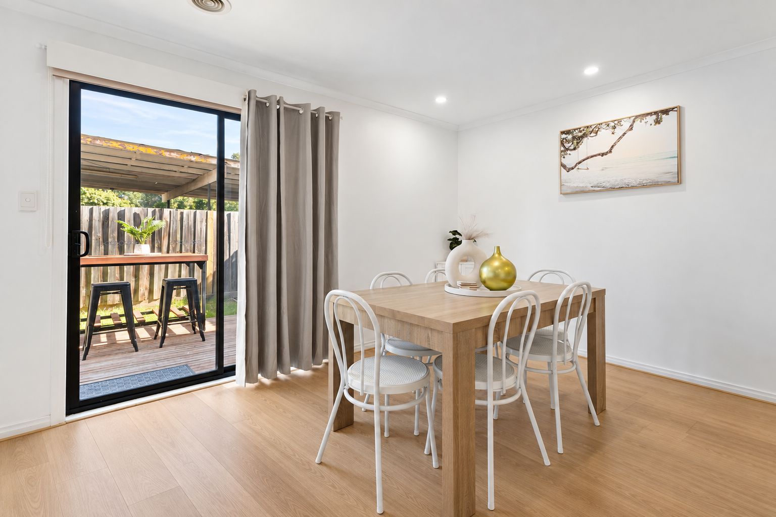 A dining area with a wooden table and white chairs, a sliding glass door leading to an outdoor deck, and a framed artwork on the wall.