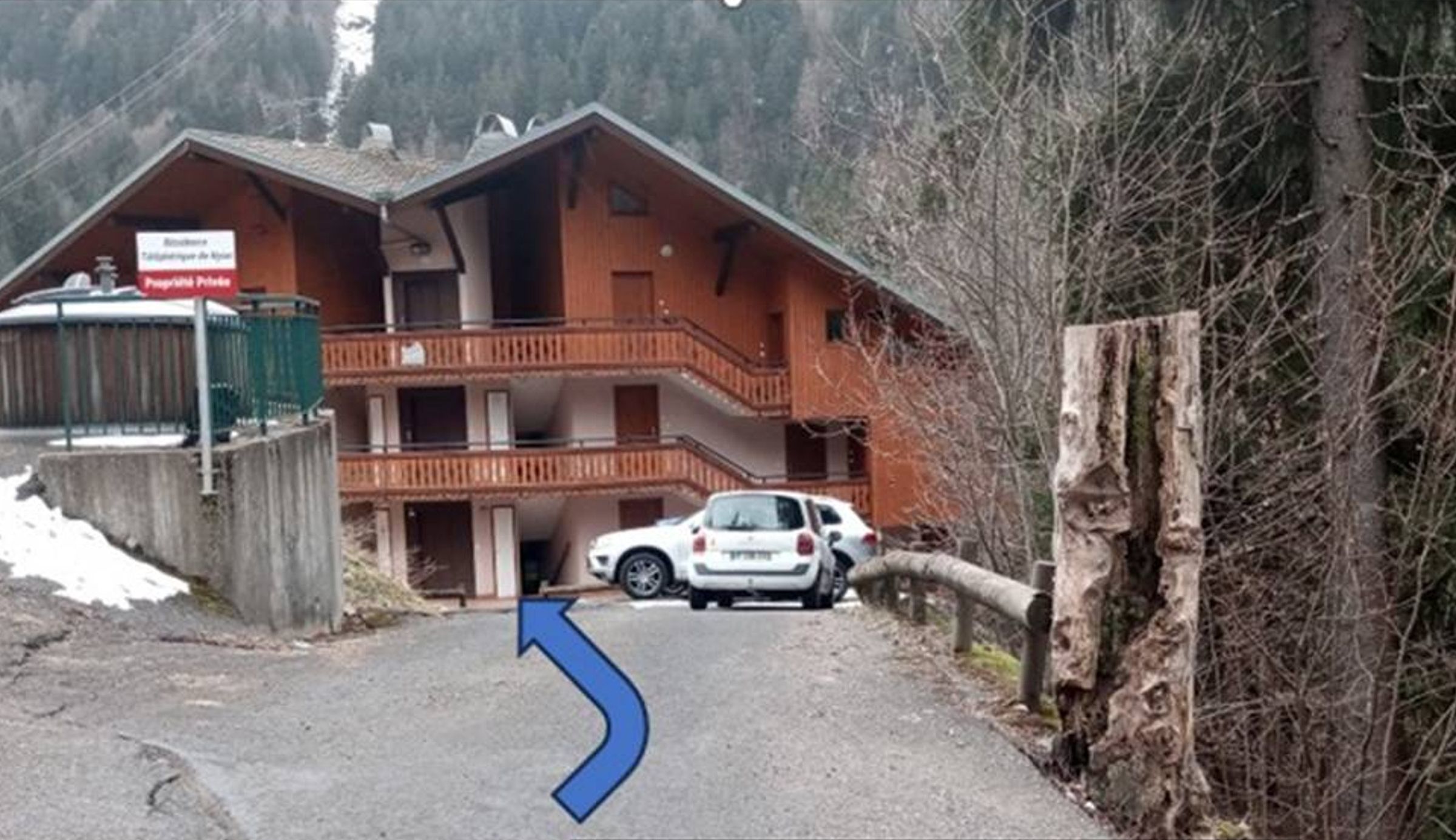 Un edificio de madera con balcón, un coche estacionado en frente y un letrero que indica 'Entrée résidence CHAMOISIERE-BAT du milieu'.