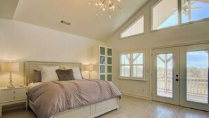A spacious bedroom with a large bed, a nightstand, a dresser, and a walk-in closet. There are large windows and a balcony.