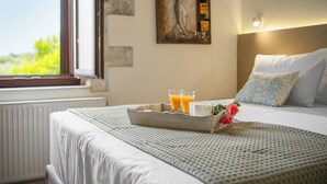 A neatly made bed with a tray of orange juice and a rose, a painting on the wall, and a window with a view of greenery. - The Olive Mill House Luxe Villa (Rethymno)