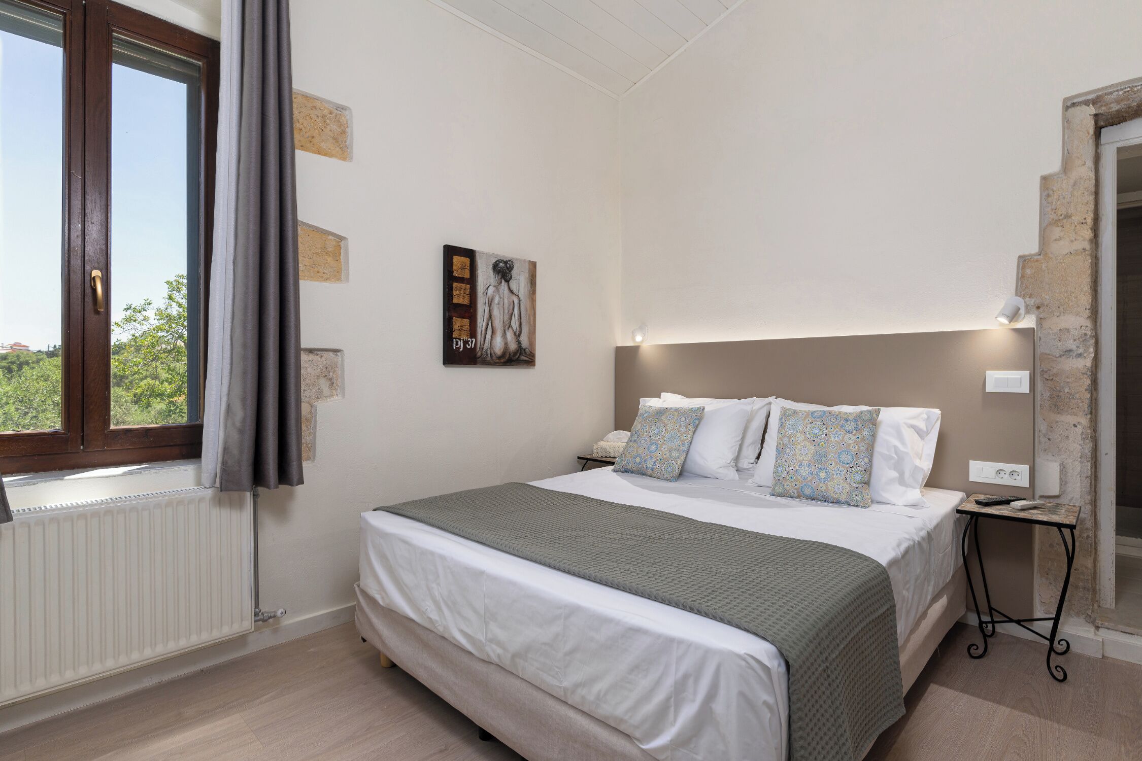 Quarto com cama, mesa de cabeceira, janela com cortinas, radiador e uma obra de arte pendurada na parede.