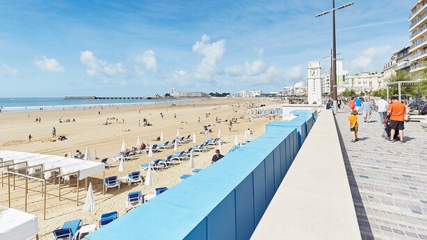 Een strand met ligbedden en parasols, een promenade met mensen en een pier op de achtergrond.