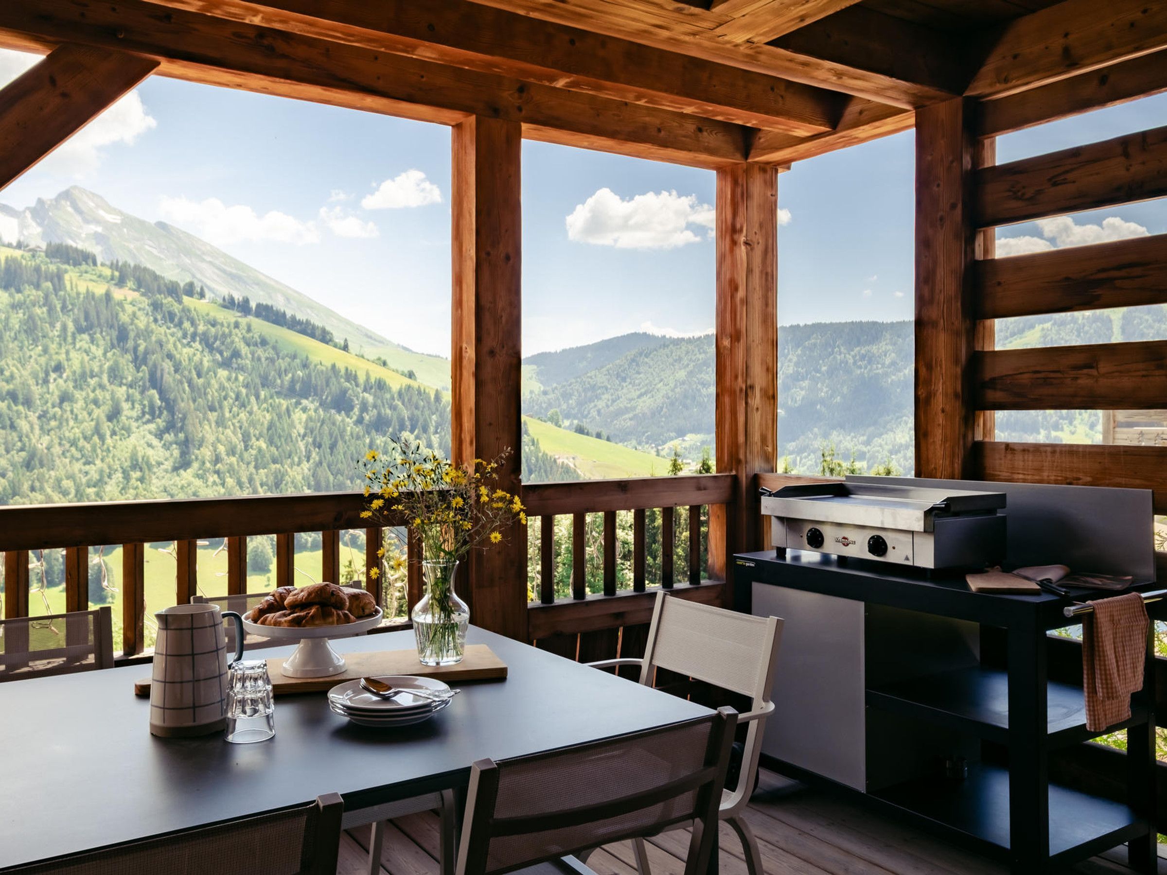 A wooden deck with a table set for breakfast, a grill, and a view of a mountainous landscape.